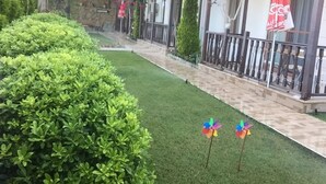 A hotel courtyard with neatly trimmed hedges, a paved walkway, and colorful pinwheels. - Gaye Apart (Ula)