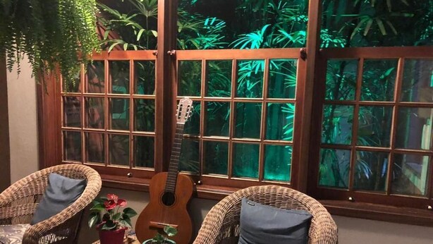 A room with wicker chairs, a guitar, and potted plants near large windows with a view of greenery. - Pousada Brisa de Paraty (Paraty)