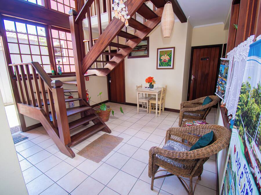 Una escalera de madera que conduce a un segundo piso, un área de comedor con sillas de mimbre, una mesa con un jarrón de flores y una pared con obras de arte enmarcadas.