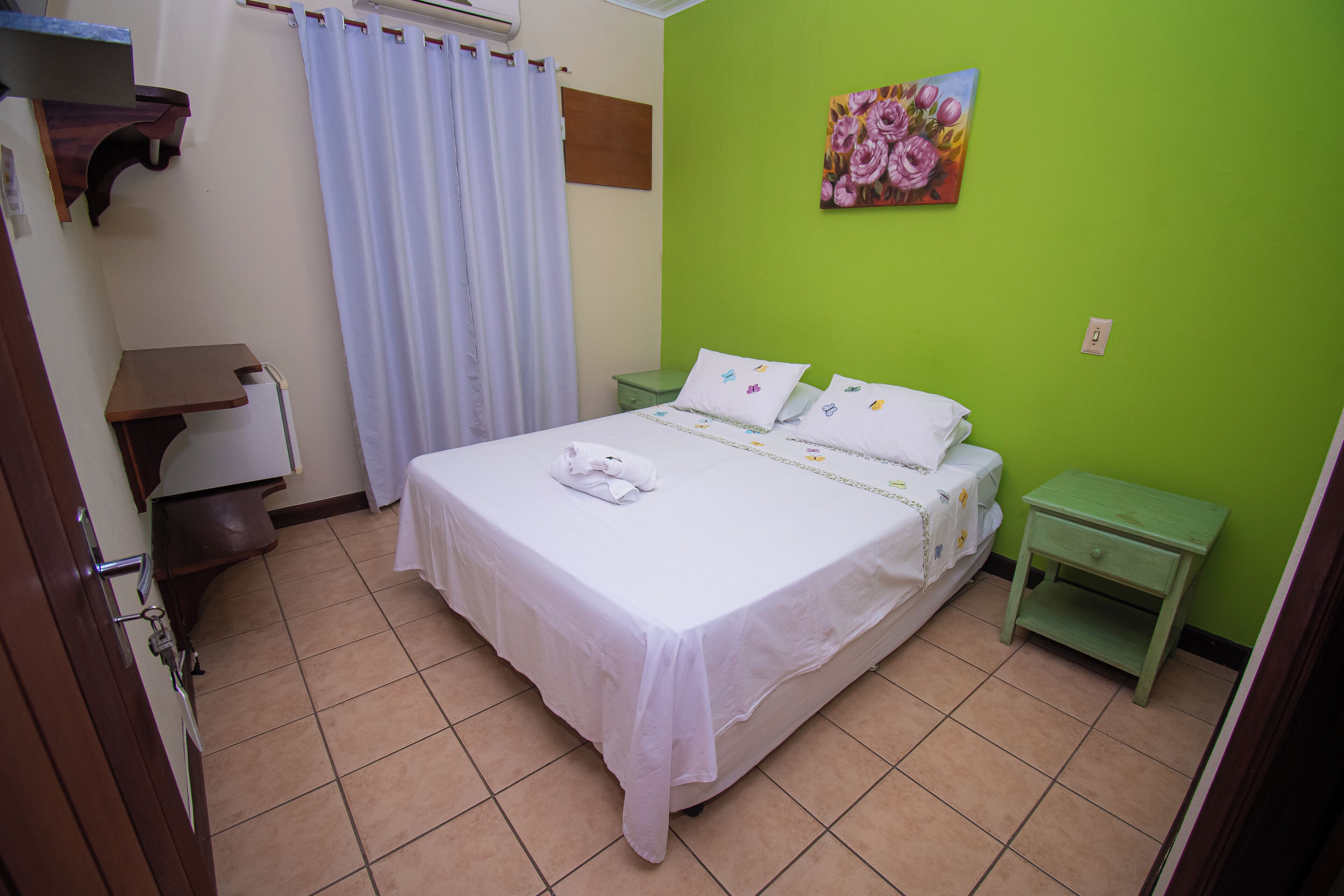Quarto bem arrumado com cama, parede verde, um quadro e uma pequena mesa de cabeceira.