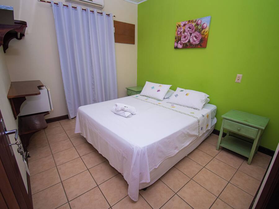 Quarto bem arrumado com cama, parede verde, um quadro e uma pequena mesa de cabeceira.