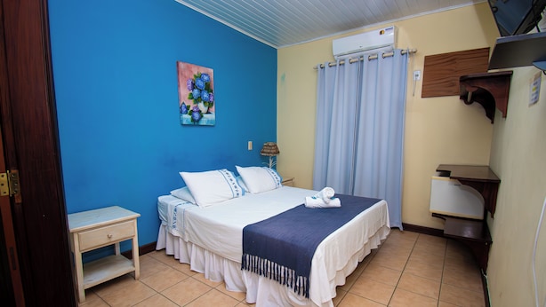 Um quarto com cama, mesa de cabeceira, quadro, aparelho de ar condicionado e janela com cortinas.