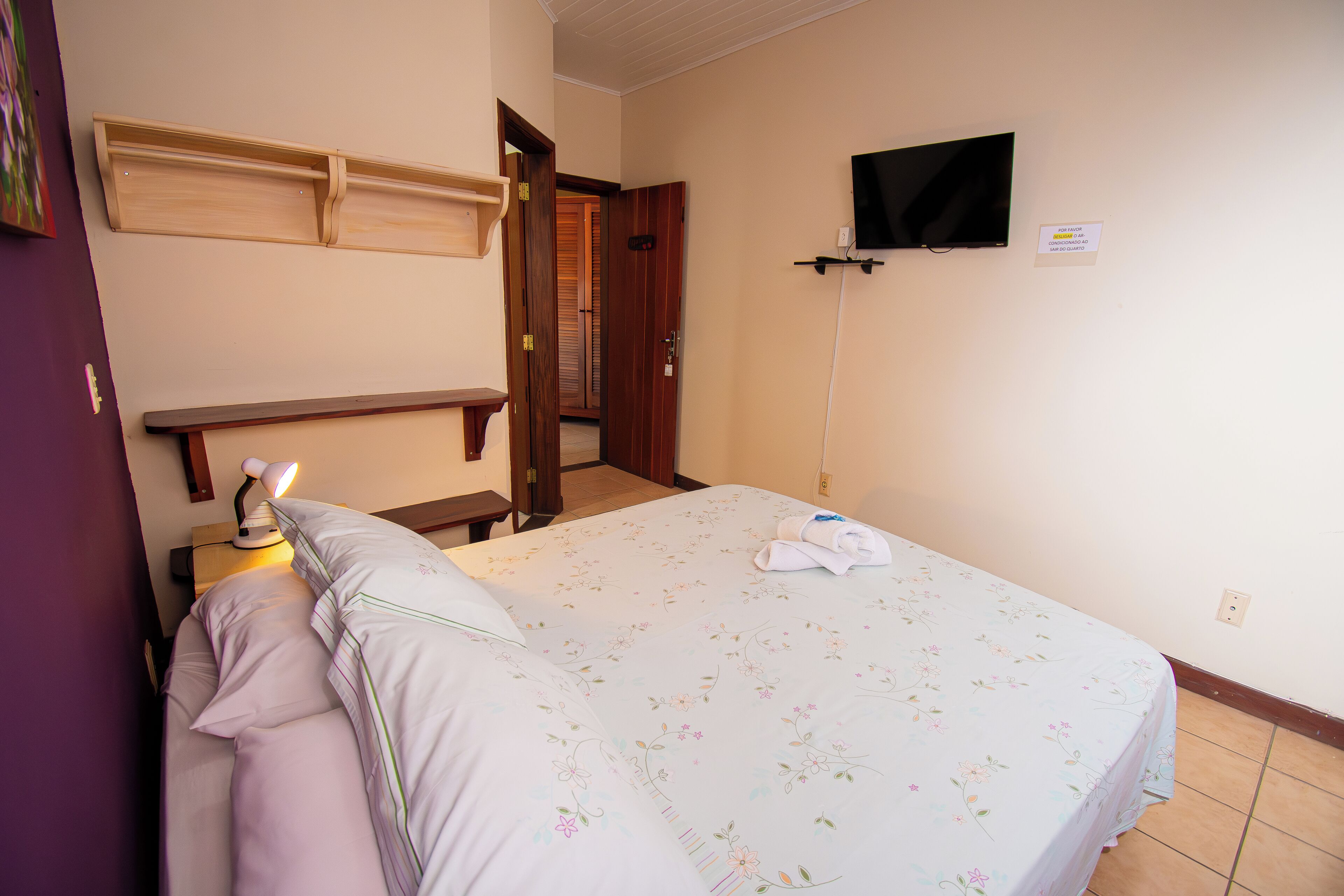 A bedroom with a bed, a nightstand, a desk, a television, and a door leading to another room.