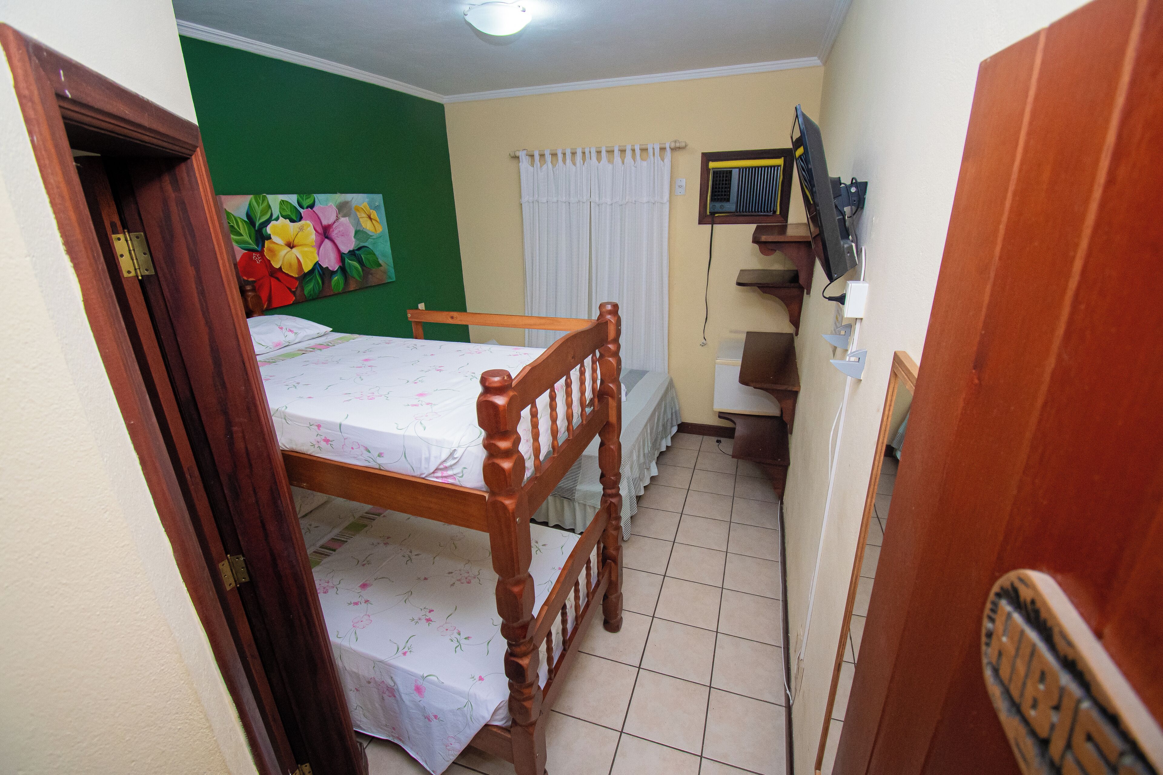 Una habitación pequeña y estrecha con una cama de madera, una pared verde, una cortina blanca y un televisor pequeño en la pared.