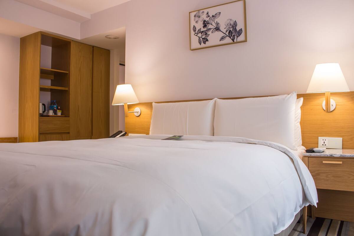 A hotel room with a large bed, wooden wardrobe, bedside lamps, and a framed floral artwork on the wall.