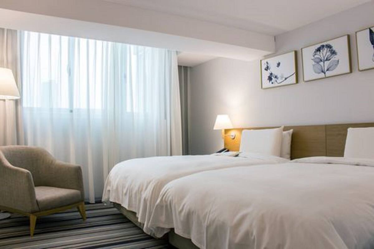 A hotel room with two beds, a chair, a lamp, and a window with sheer curtains.