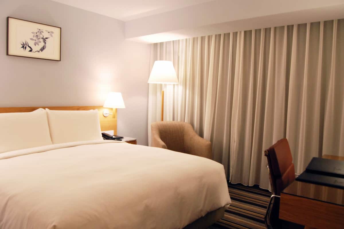 A hotel room with a bed, a chair, a desk, a lamp, a nightstand, and a curtain.