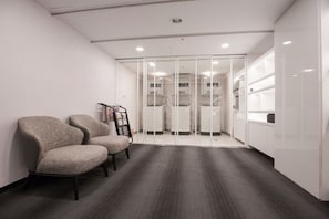 Un espacio de oficina moderno con dos sillones tapizados, una pizarra y un separador de vidrio con vista a una zona de cocina.
