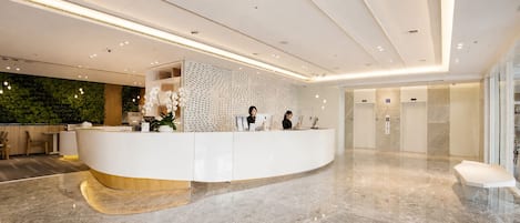 Un área de recepción moderna con un mostrador curvo, dos recepcionistas, una pared verde con plantas y un piso de mármol.