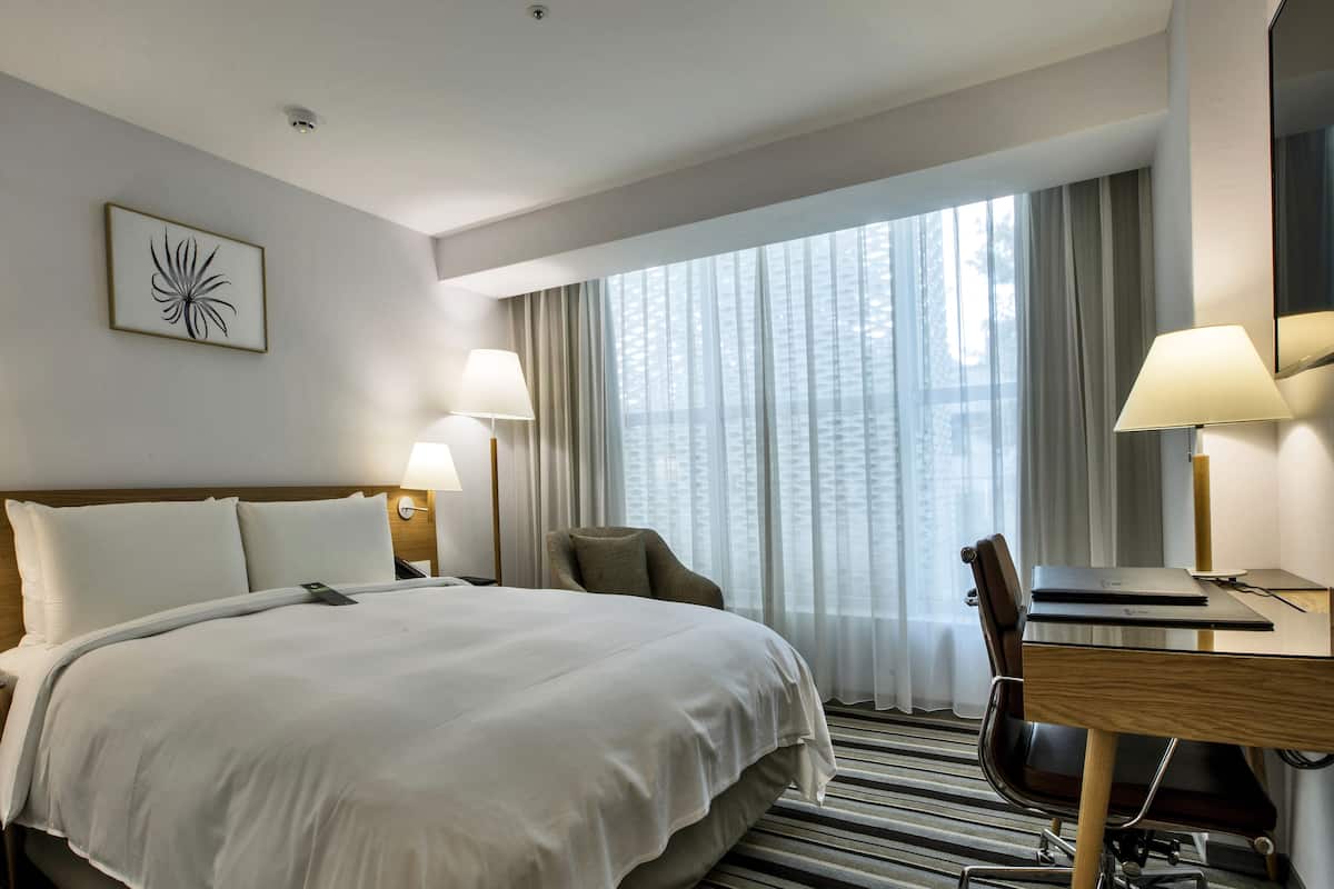 A hotel room with a large bed, a desk with a chair, a lamp, and a window with curtains.