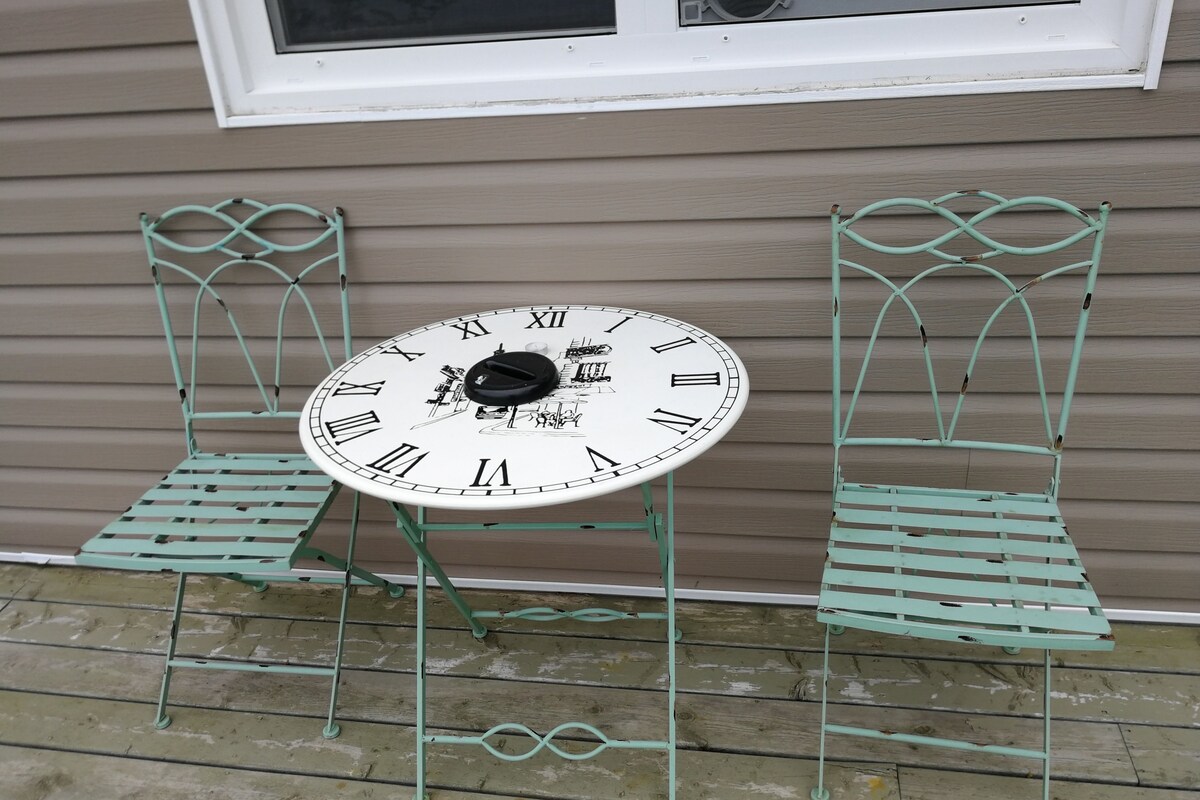 A round table with a clock face, two metal chairs, and a window in the background.