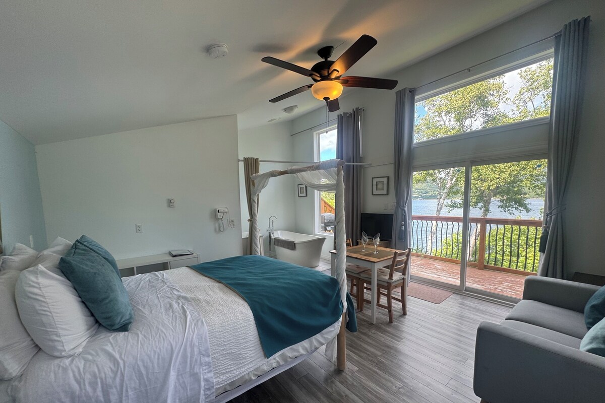 A bedroom with a bed, a desk, a chair, a balcony with a view of the water, and a ceiling fan.