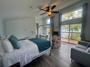 A bedroom with a bed, a desk, a chair, a balcony with a view of the water, and a ceiling fan. - Knotty Pine Cottages (Ingonish Ferry)
