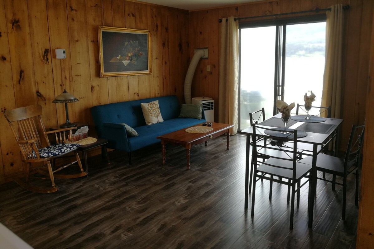 A cozy room with a blue sofa, wooden flooring, a dining table with chairs, a rocking chair, and a painting on the wall.