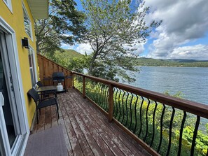 A wooden deck with a grill and chairs, overlooking a lake with a forested shoreline. - Knotty Pine Cottages (Ingonish Ferry)