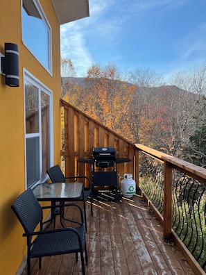 A wooden deck with a grill, table, and chairs, overlooking a forested area. - Knotty Pine Cottages (Ingonish Ferry)