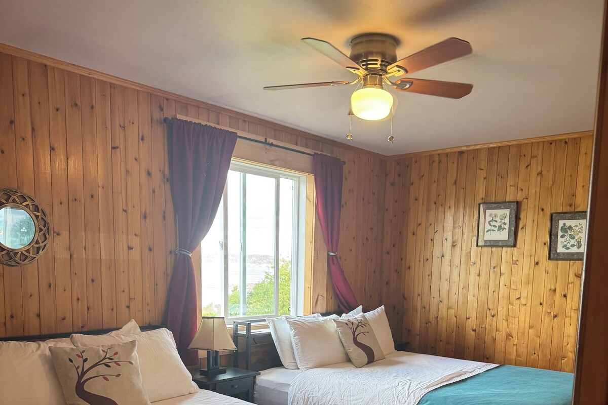 A room with two beds, wooden walls, a ceiling fan, and a window with curtains.