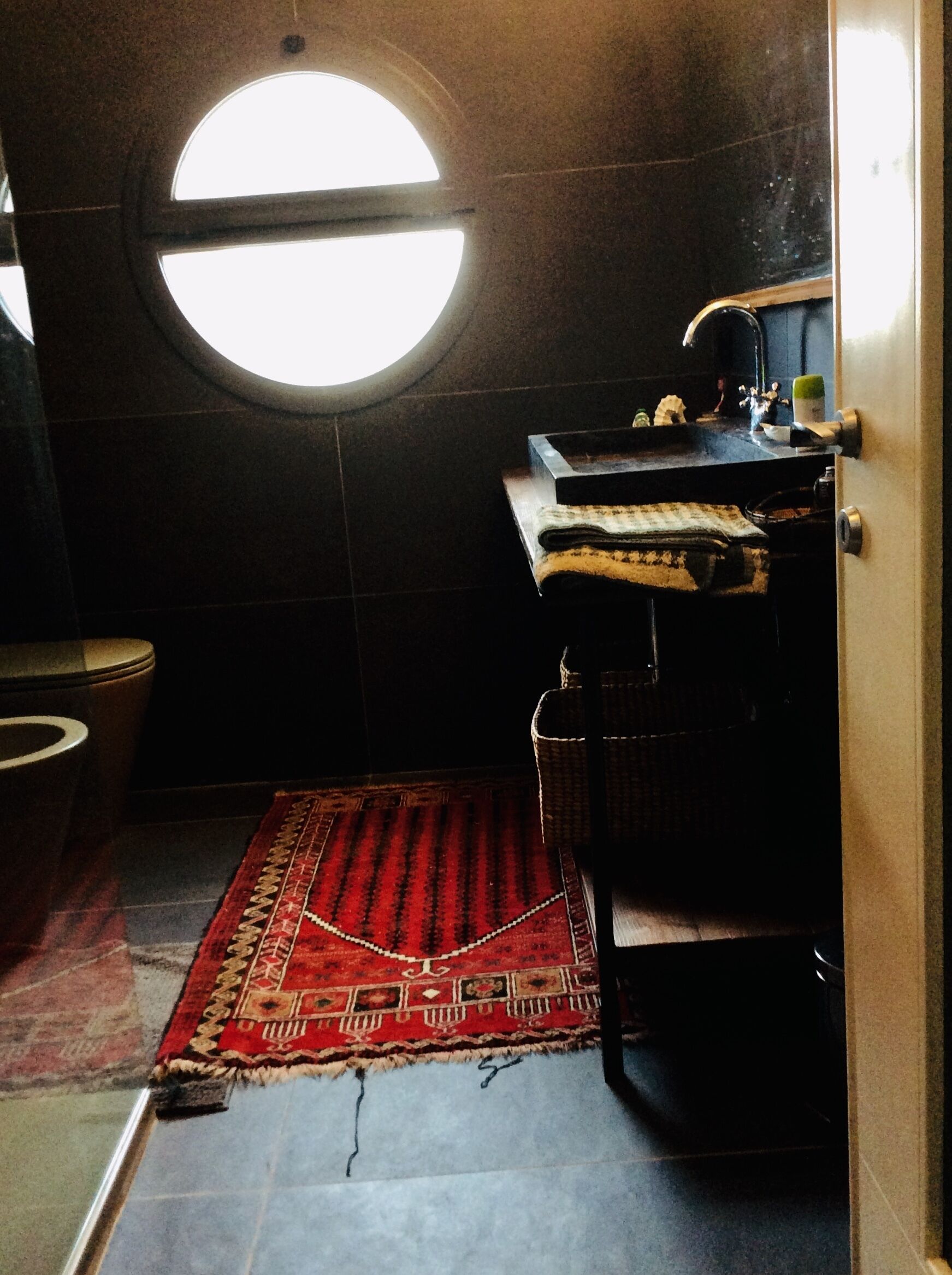 Een badkamer met een rond raam, een toilet, een wastafel en een vloerkleed.