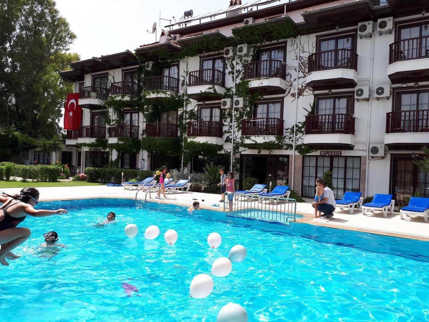 A swimming pool with people and white balloons, surrounded by a building with balconies and air conditioning units.
