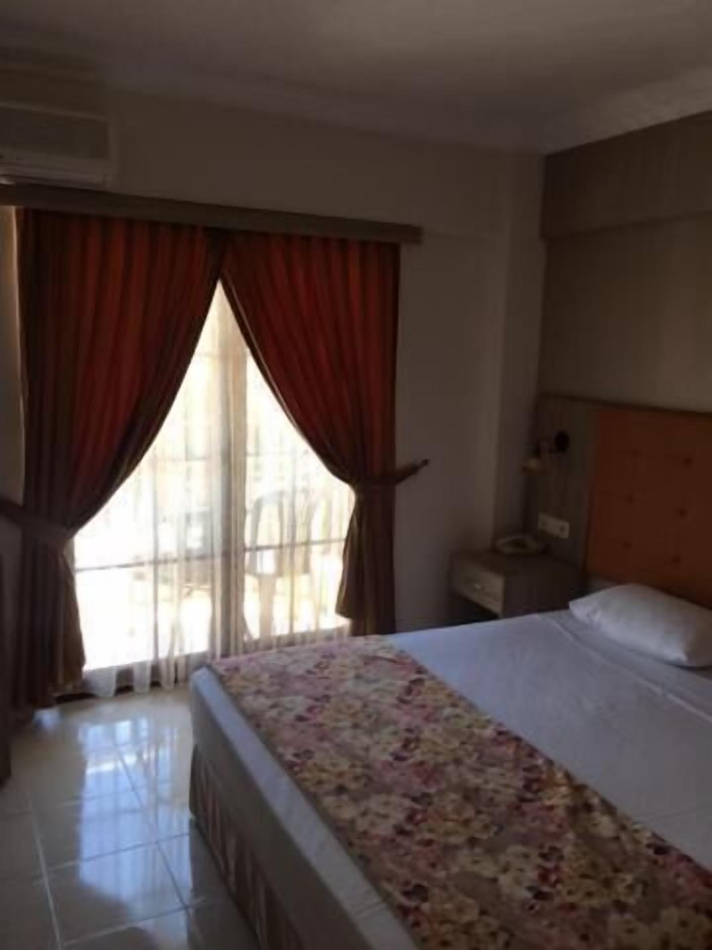 Une chambre d’hôtel avec un lit, une table de chevet, une fenêtre avec des rideaux et un sol carrelé.