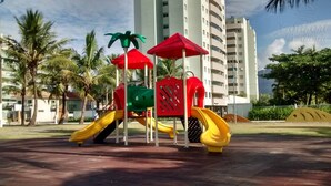A playground with slides and climbing structures, surrounded by palm trees and a tall building in the background. - Doce Lar de Bertioga (Bertioga)