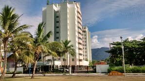 A high-rise building with multiple balconies, surrounded by palm trees and a mountainous backdrop. - Doce Lar de Bertioga (Bertioga)