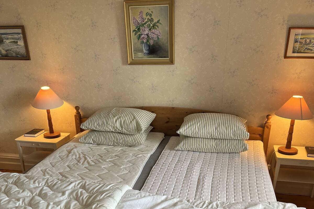 Una cama doble con edredón blanco acolchado, dos mesitas de noche con lámparas, almohadas a rayas y un cuadro enmarcado en la pared.