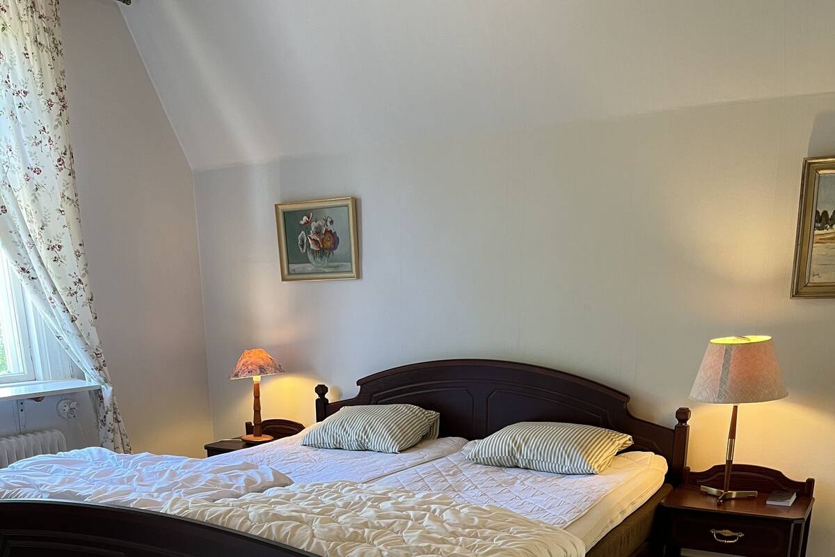 Un dormitorio con una cama de madera oscura, ropa de cama blanca, dos mesitas de noche con lámparas, una ventana con cortinas florales y un cuadro en la pared.