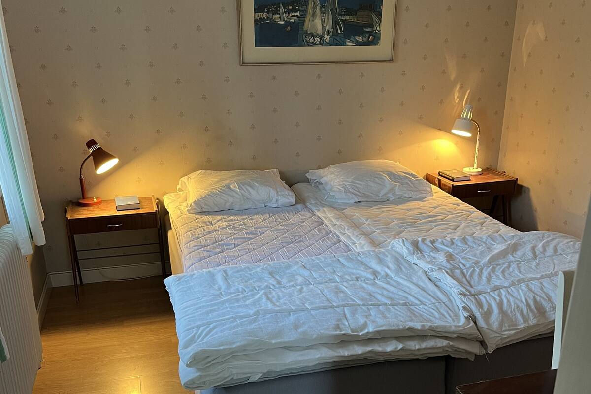 Una cama bien hecha con sábanas blancas, dos mesitas de noche con lámparas, un cuadro enmarcado en la pared y suelo de madera.