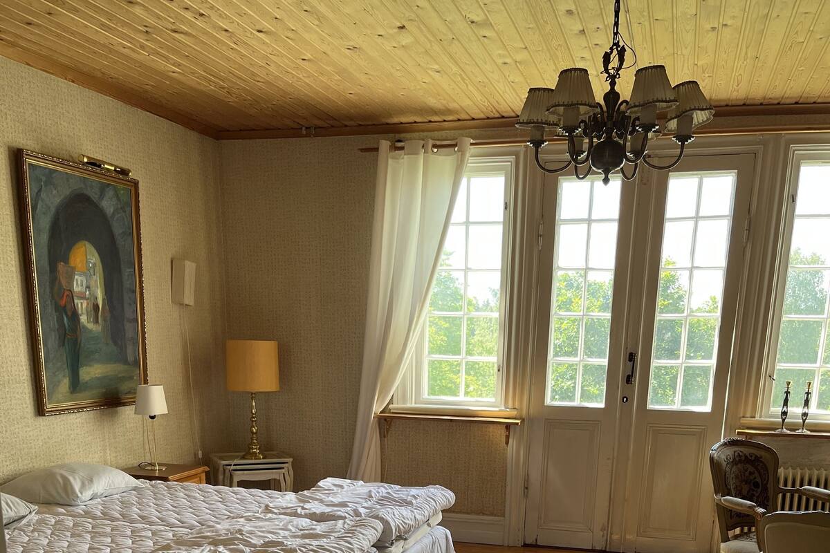 Un dormitorio con una cama grande, un candelabro, un cuadro y una ventana con cortinas.