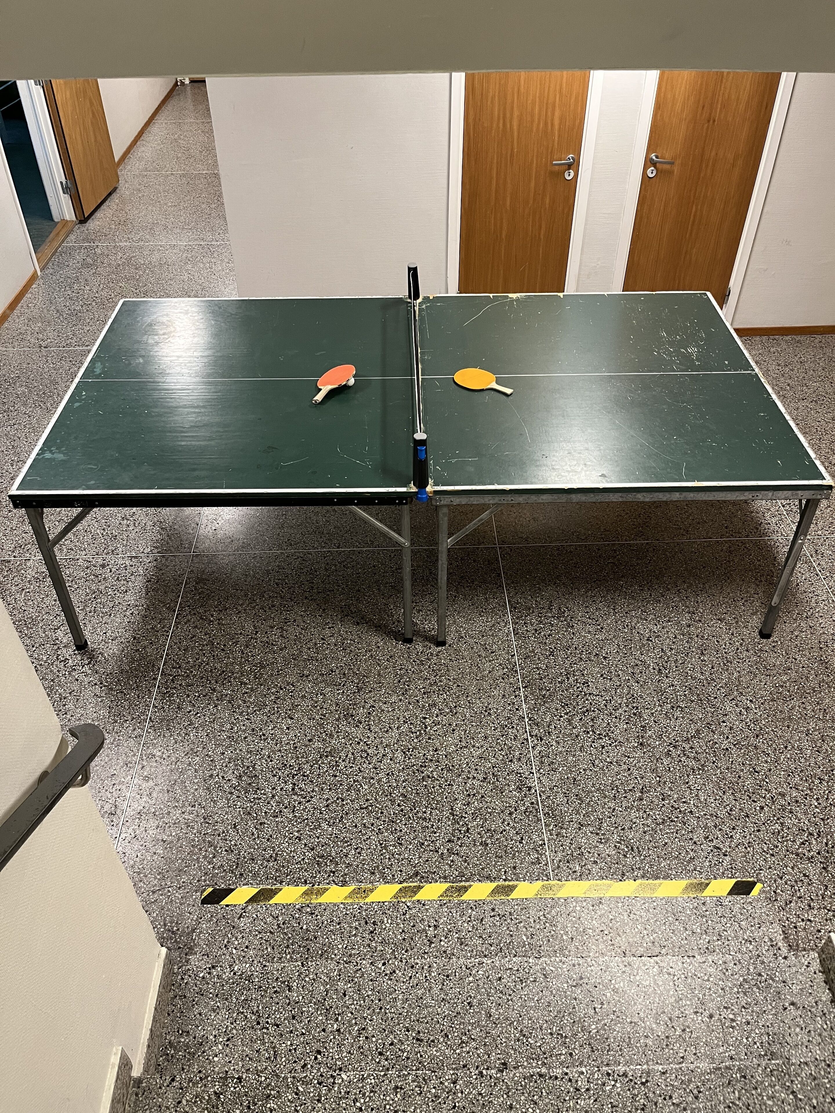 A ping pong table with two paddles in a room with a tiled floor and a yellow caution tape marking an area.
