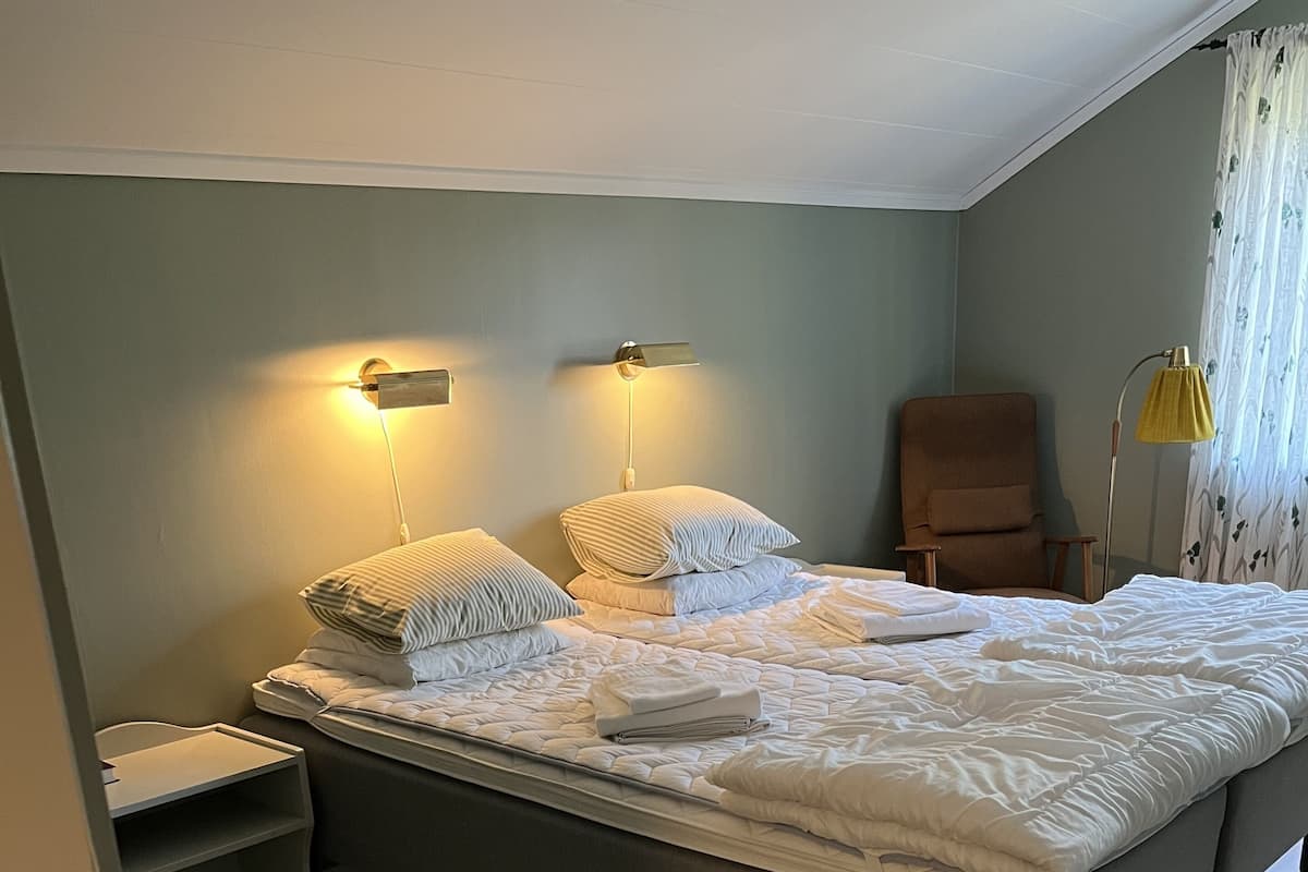 Una cama bien hecha con almohadas, una mesita de noche, una silla y una lámpara de pie en una habitación con suelo de madera y paredes de color claro.
