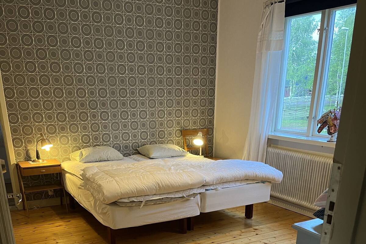 Un dormitorio con suelo de madera, cama con ropa de cama blanca, mesita de noche con lámpara y papel tapiz estampado.