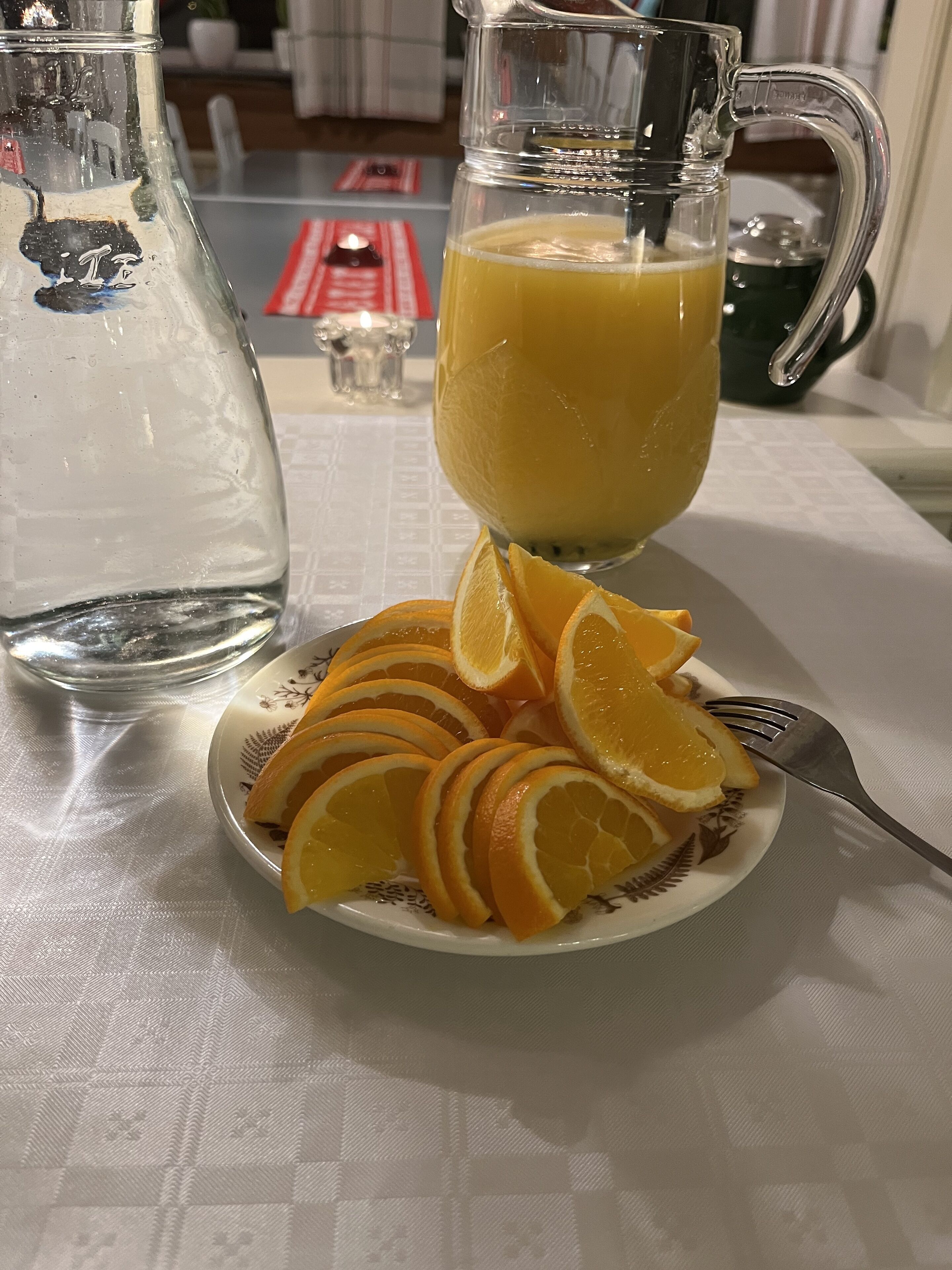 En glasbringa med apelsinjuice, ett glas vatten och en tallrik med apelsinskivor står på ett bord.