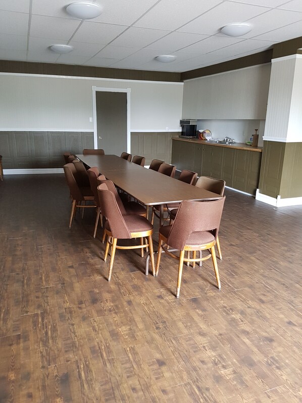 A conference room with a long table and chairs, wooden flooring, and a kitchenette in the background. - Seaport Inn (Trinity Bay North)