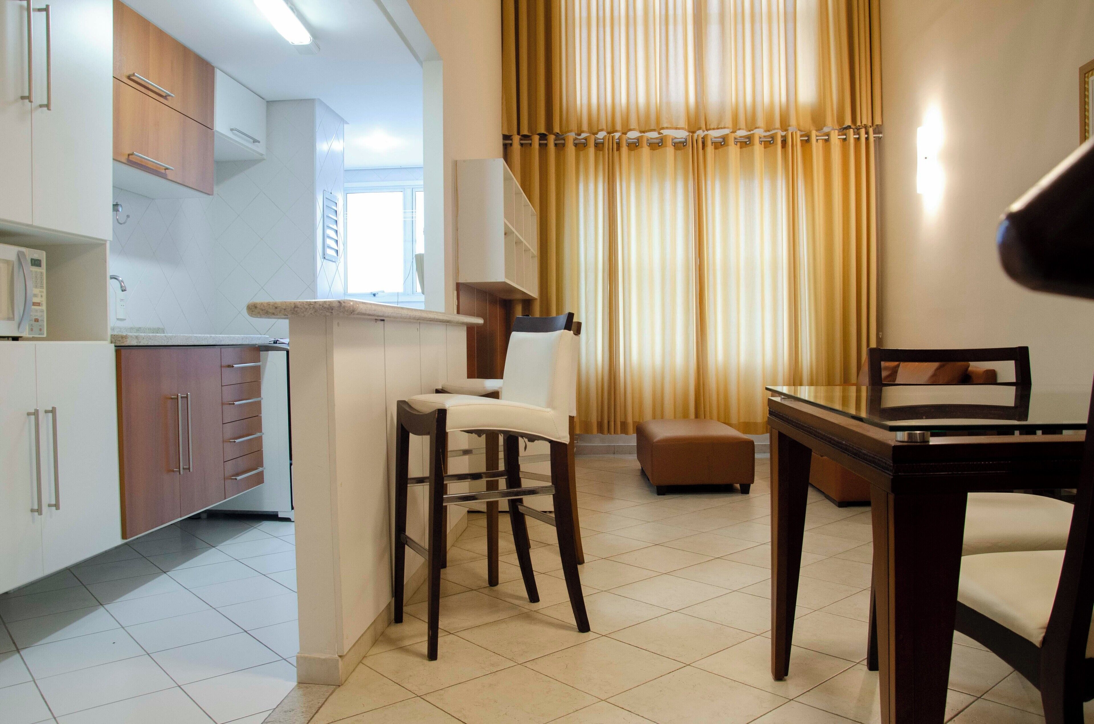 A modern kitchen with white cabinets, a built-in microwave, and a small island with stools. There is a dining area with a wooden table and chairs, and a large window with curtains.