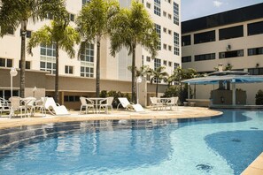 A swimming pool with lounge chairs and tables, surrounded by palm trees and a building with multiple windows. - Hotel City Hall (São José dos Campos)