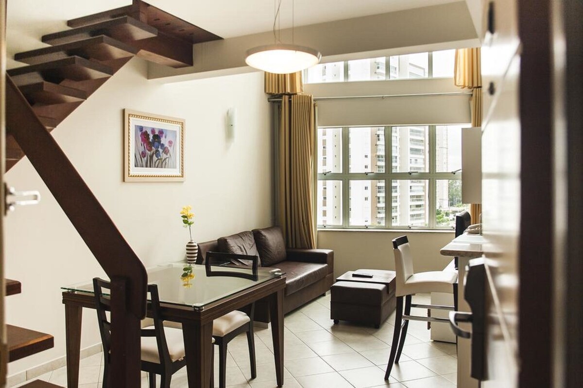 A modern living room with a staircase, a dining area, a sofa, and a large window with curtains.
