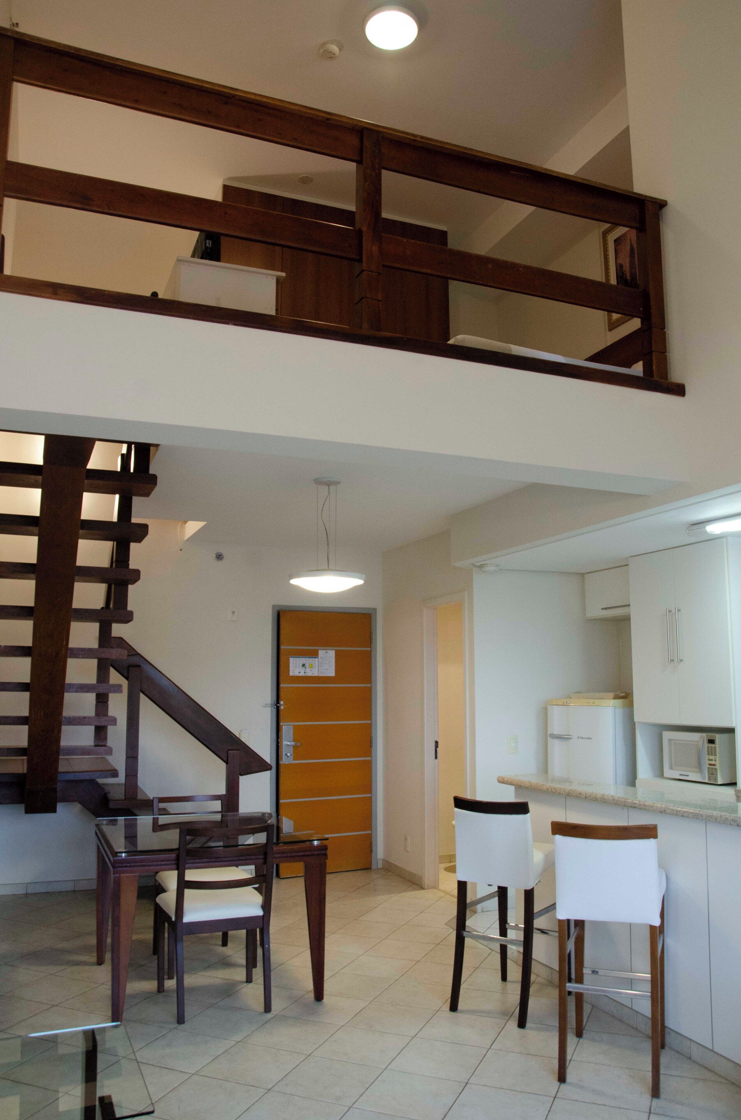 Un comedor con mesa de vidrio y sillas de madera, una cocina con armarios blancos y un microondas, y una escalera de madera que conduce a un nivel superior.