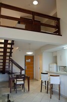 Un comedor con mesa de vidrio y sillas de madera, una cocina con armarios blancos y un microondas, y una escalera de madera que conduce a un nivel superior.