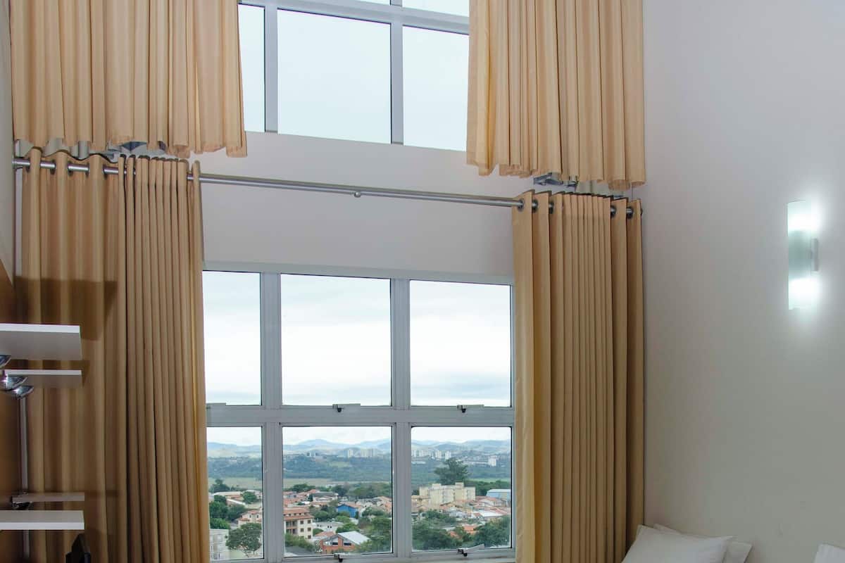 A hotel room with two beds, a desk, a chair, and a view of a cityscape through large windows.