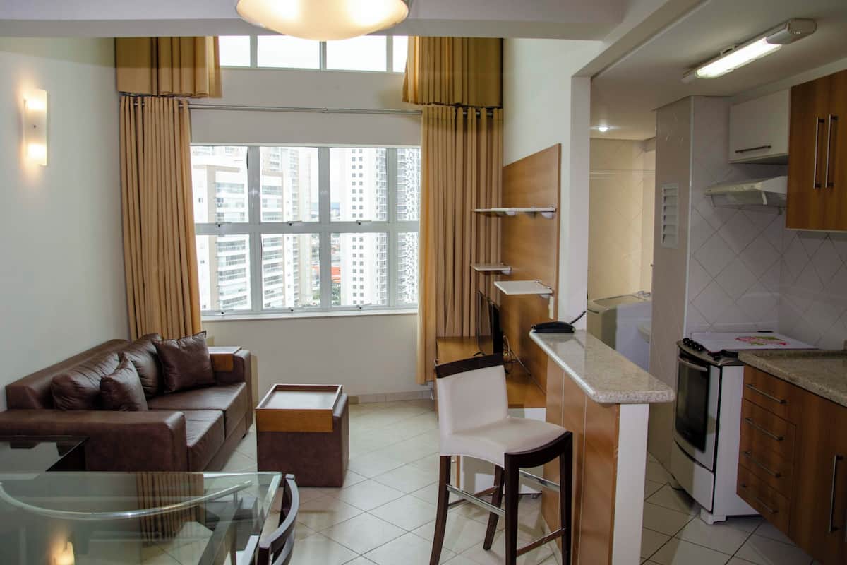 A compact living space with a sofa, a dining table, a kitchen area including a stove, and a window with curtains.