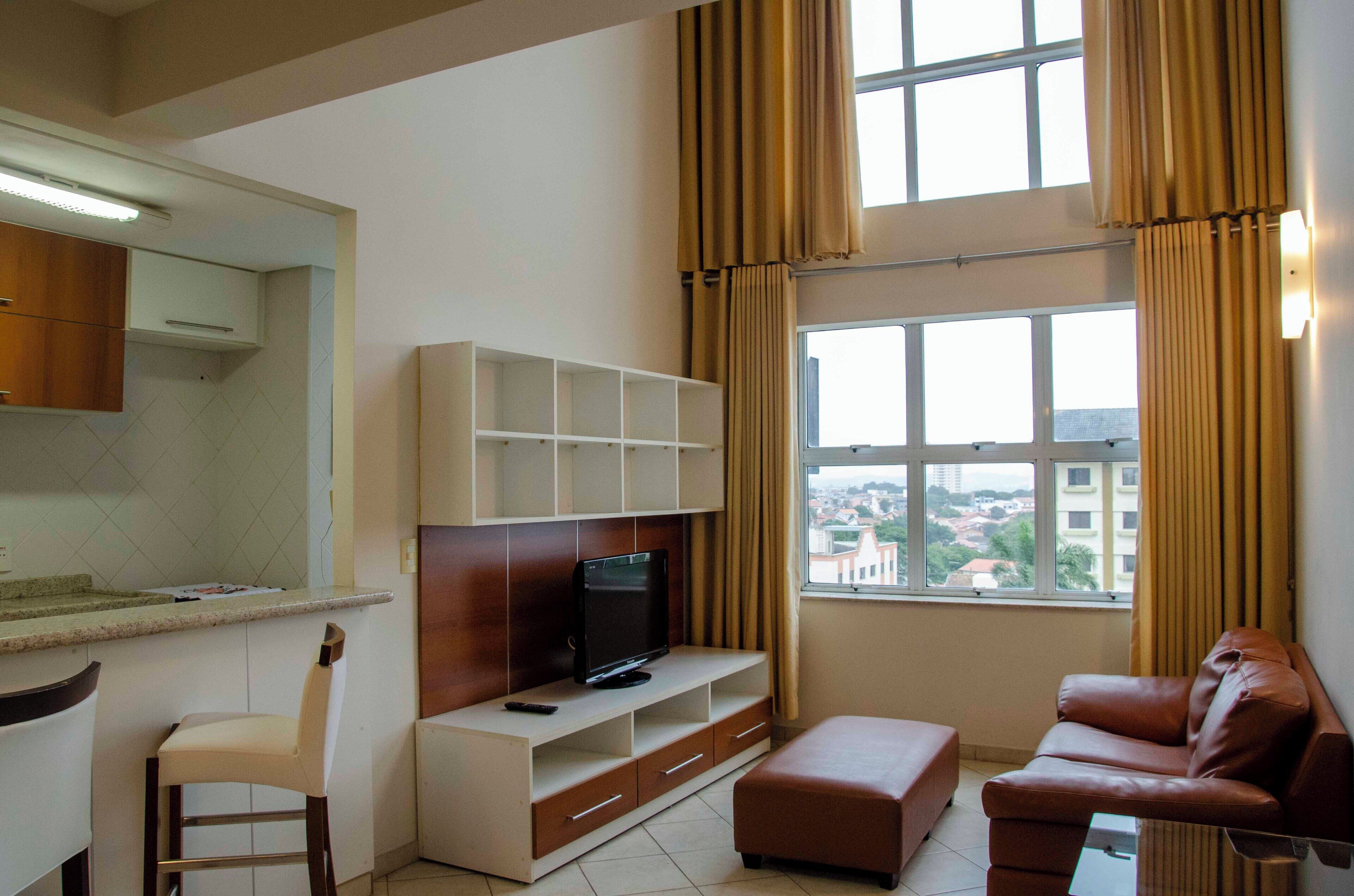 Una sala de estar moderna con televisor, sofá, zona de comedor con mostrador y un amplio ventanal con cortinas.
