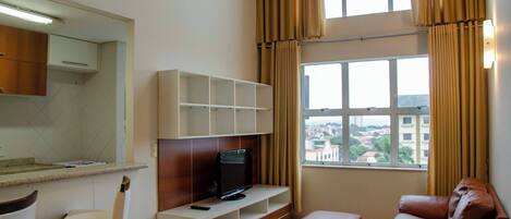 Una sala de estar moderna con televisor, sofá, zona de comedor con mostrador y un amplio ventanal con cortinas.