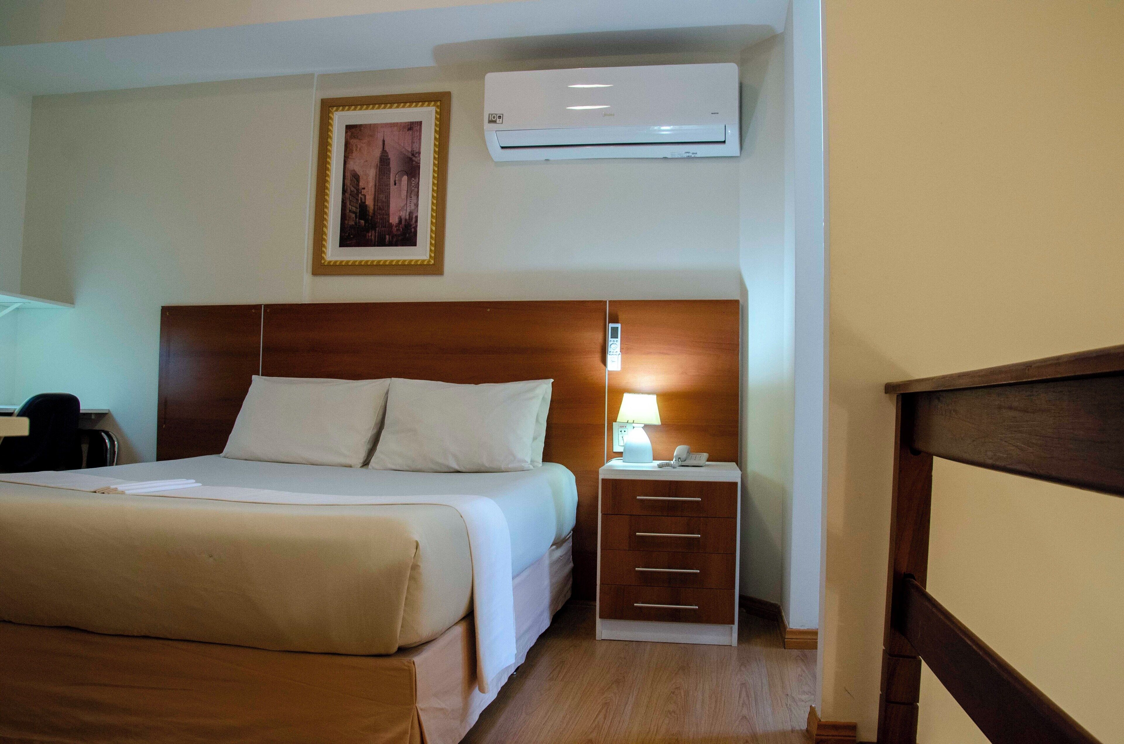 Quarto de hotel com cama, luminária de cabeceira, mesa de apoio e aparelho de ar condicionado.