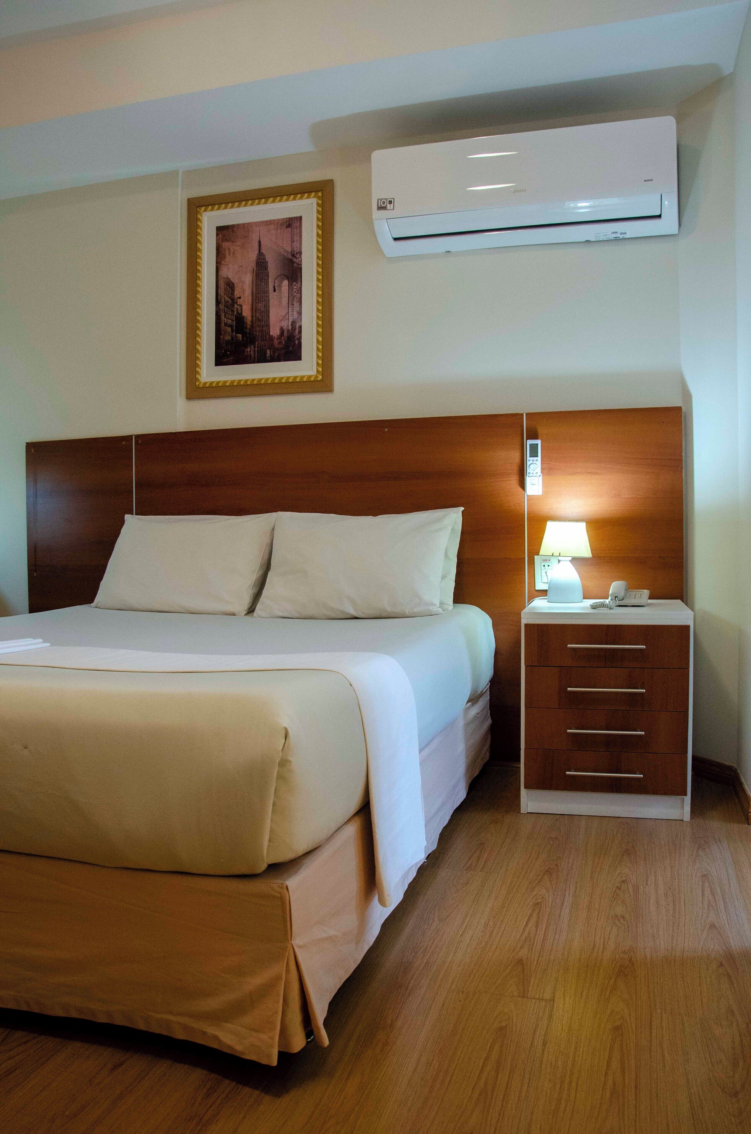 A hotel room with a wooden headboard, a bed with white linens, an air conditioning unit, a bedside lamp, and a framed picture on the wall.