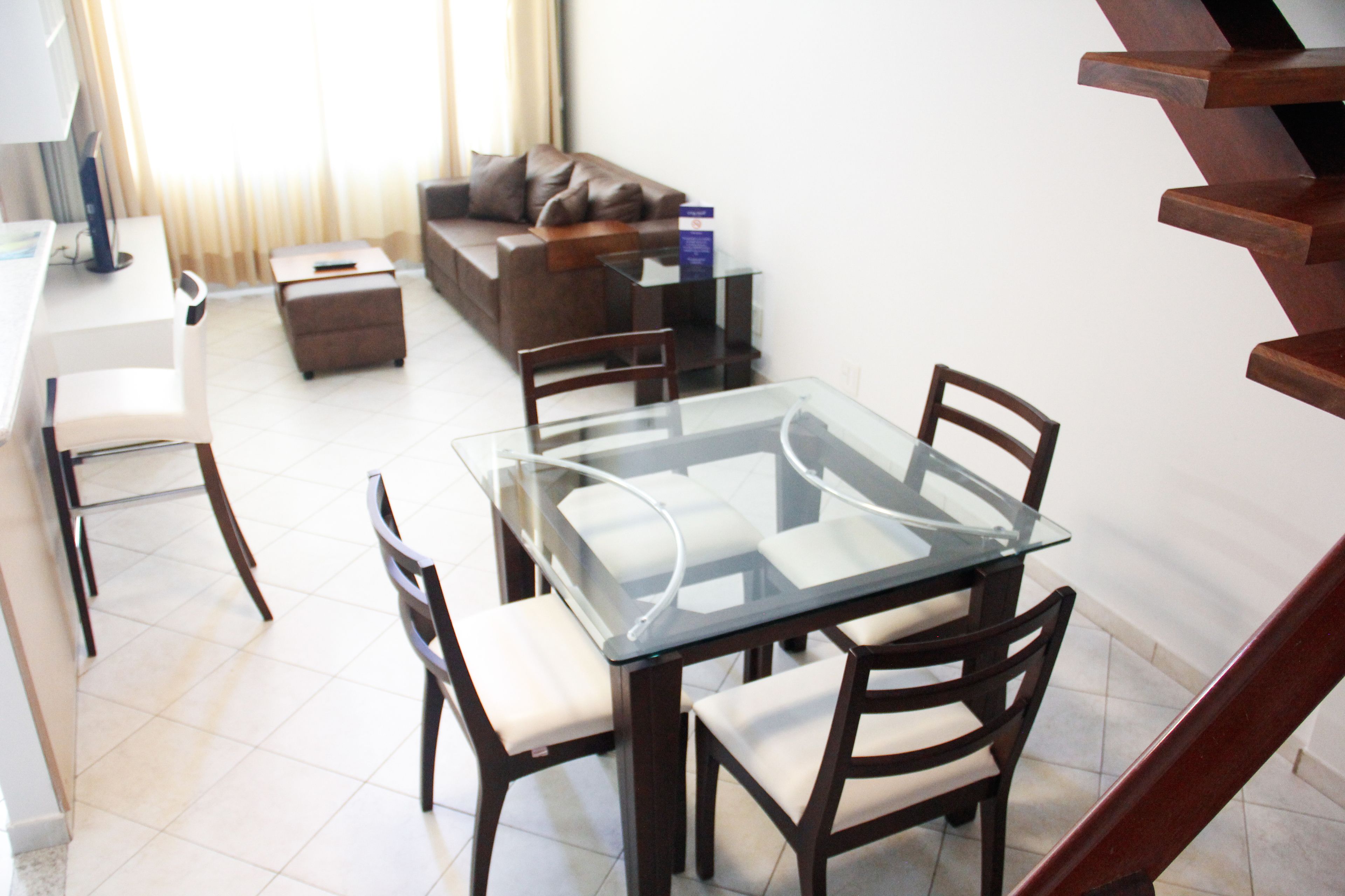 A modern living room with a glass-top dining table, wooden chairs, a brown leather sofa, and a small side table.