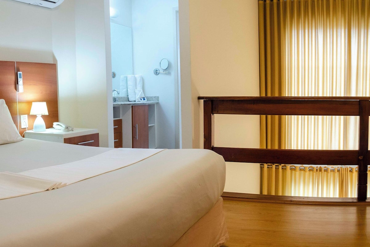 A hotel room with a wooden headboard, a bed with white linens, a bedside table with a lamp, a small bathroom with a mirror, and a window with curtains.