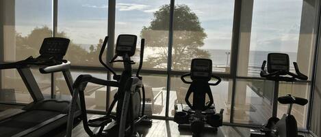 A gym with treadmills, elliptical machines, and stationary bikes, with large windows offering a view of the outdoors.