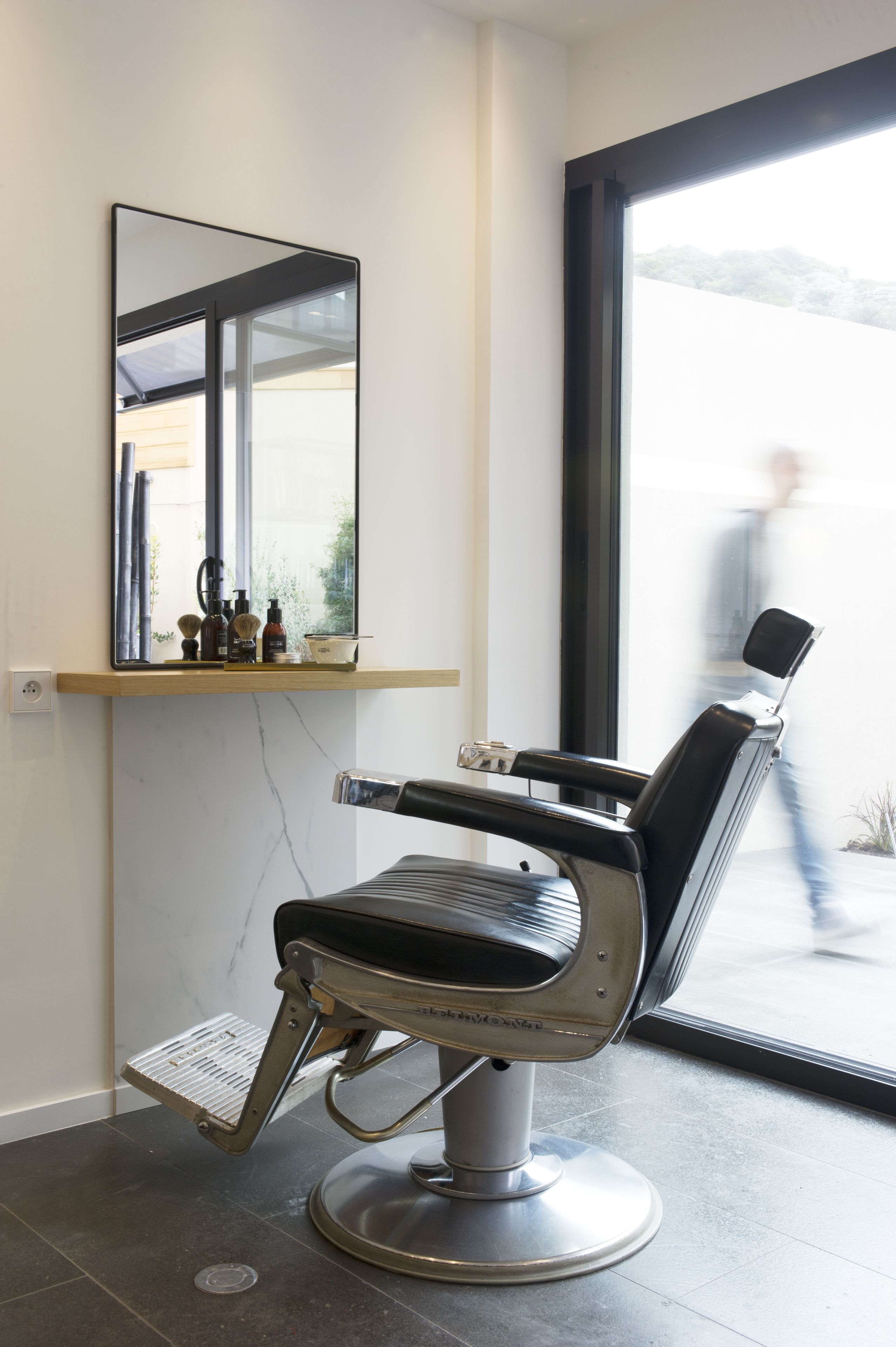 A modern barber chair with a mirror above it, a shelf with grooming products, and a view of a street outside.